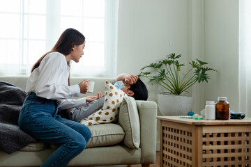 A woman sits on a couch next to a man who is sick. She is holding a cup and talking to him