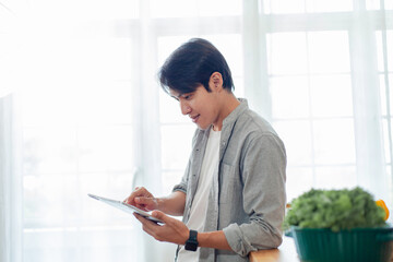 A man is sitting in front of a window, looking at a tablet. He is smiling and he is enjoying himself