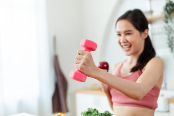 A woman is holding a pink dumbbell and smiling. She is in a kitchen with a bowl of vegetables on...
