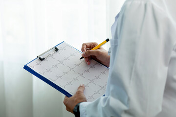 A doctor is writing on a clipboard with a yellow pen. The clipboard has a heart on it