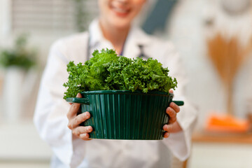 A woman wearing a white lab coat is holding a green container with a bunch of greens in it