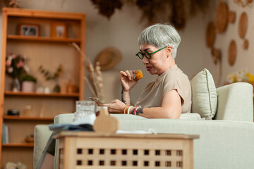 A woman is sitting on a couch and eating a snack. She is looking at her phone. The room has a lot of decorations, including a vase and a potted plant