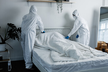 Two people in white suits are cleaning a bed. The bed is covered in a white sheet and a blanket