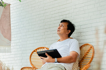 A man is sitting on a wicker chair with a tablet in his hand. He is focused on the screen, possibly...