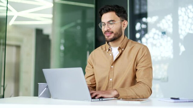 Handsome entrepreneur working typing on a laptop sitting at a workplace in a business office. Happy businessman received good news on computer. Smiling glad man worker chatting online with a client