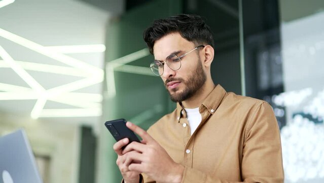 Serious handsome businessman uses mobile phone sitting at workplace in a business office. Man worker is working on application, chatting with client, banking or ordering online on smartphone. Close up