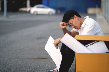 A man sits on the ground with a box of papers in front of him. He looks upset and frustrated
