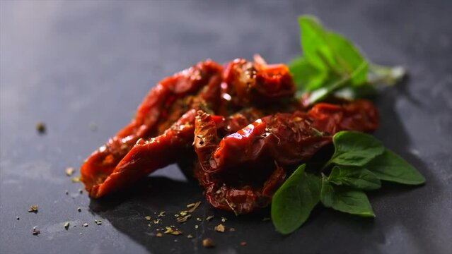 Sun dried tomatoes with aromatic herbs, oregano, marjoram leaf. Rotating on black stone background, close up. Italian cuisine, healthy vegan food, Mediterranean diet. Dry tomato. 