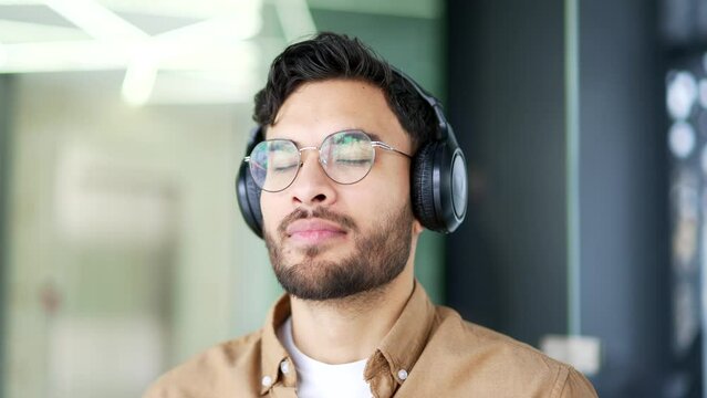 Happy businessman in wireless headphones relaxing with his eyes closed sitting at workplace in a business office. Calm man listening to relaxing music enjoys sounds of relaxation and noise of nature