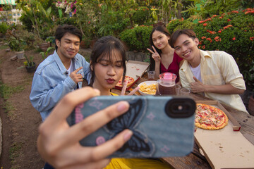 A young asian woman takes a selfie together with three of her friends while enjoying pizza and other snacks during a picnic at the lawn.