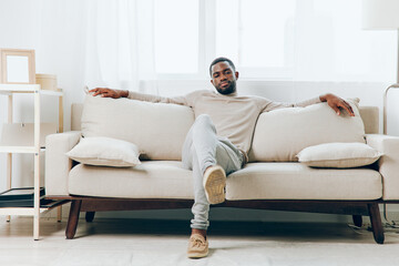 Sad African American man sitting on a comfortable sofa in his modern living room, deeply lost in...
