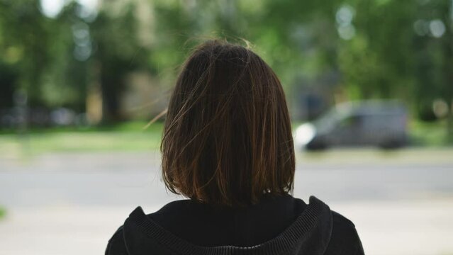 Rear view of adult brunette female standing on the street and looking at traffic and cars passing by