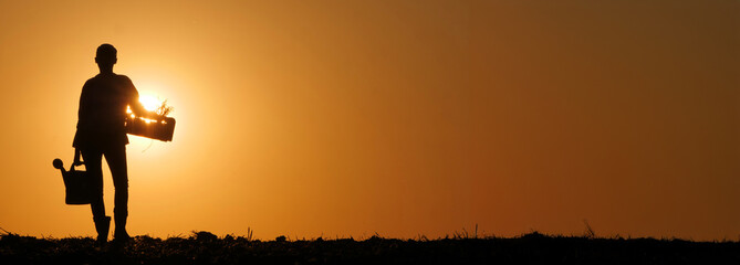 At sunset, the outline of a woman farmer is visible in a field, clutching a watering can and a...