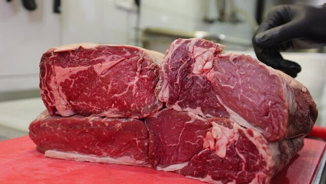 Experienced butcher in black sterile gloves squeezing four pieces of marbled beef and placing on red board in kitchen. Caucasian man checking quality of red meat before cooking nourishment dish.