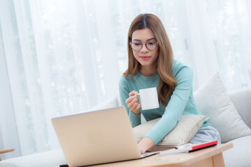 Young asian woman wearing glasses sitting sofa working on laptop computer and drinking coffee in...