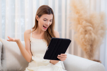 Young asian woman sitting on sofa using digital tablet computer for video call in living room at...