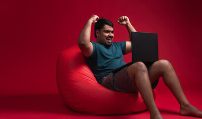 Excited person looking at laptop, Happy Indian man sitting on red chair cheering with laptop...