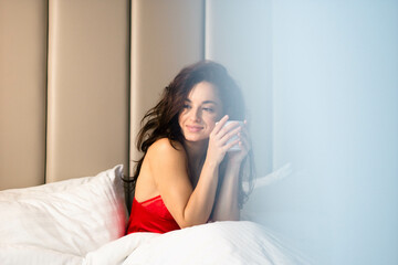 Caucasian beautiful brunette female sitting in bed in red sleepwear holding cup of coffee. Contented pretty woman in sexy red lingerie drinking morning coffee in bed. Valentine's day