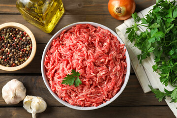 Raw ground meat in bowl, spices, oil and parsley on wooden table, flat lay