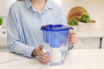 Woman with water filter jug at white marble table in kitchen, closeup. Space for text