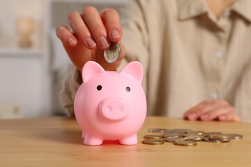 Woman putting coin into pink piggy bank at wooden table, closeup