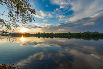 The vibrant sun dips below the horizon, casting a warm glow over the serene lake, creating a...