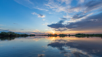 The sun sets over the water, painting the sky with a blend of colors as clouds drift by, creating a mesmerizing and serene scene.