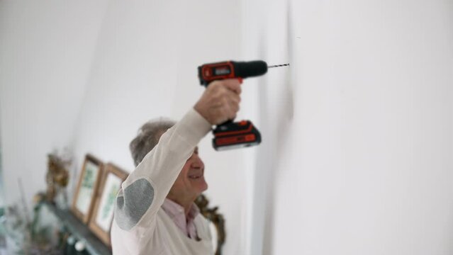 Focused elderly man using a cordless drill on a white wall, home improvement project, wearing casual clothes, determined expression, DIY task, home renovation activity