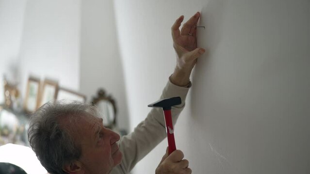 Older man hammering a nail into a white wall, concentrating on the task. DIY craftsmanship and home improvement efforts, set in a bright, cozy indoor space