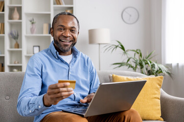 A happy man is seen shopping online with a laptop and credit card in the comfort of his living...
