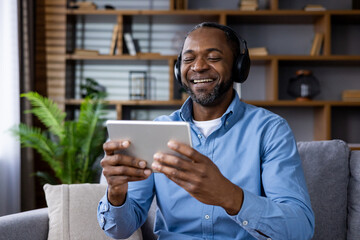 A man in a blue shirt is comfortably watching content on a digital tablet with headphones at home,...
