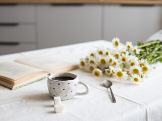A mug of coffee on a table covered with a white linen tablecloth. There is a blurred kitchen in the background. A cozy homemade breakfast. Front view. The view is horizontal.