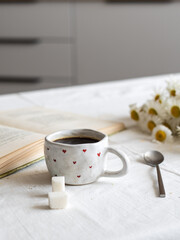 Breakfast concept, morning coffee. A cup of coffee, two pieces of sugar and an open book on a table covered with a light tablecloth against the background of the kitchen interior.
