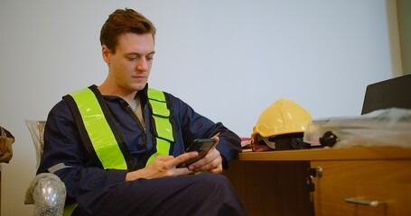 A construction worker in uniform sits in an office, taking a break and using smartphone. a moment of relaxation and communication during a workday in a construction environment