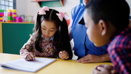 Teacher helping kid students learn to read in classroom at kindergarten school room. Multiracial and education concept. Focus on Asian toddler face