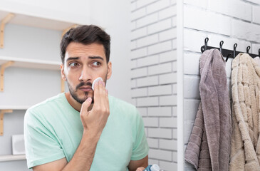 Handsome man looking at mirror and applying moisturizing cream on cheeks in bathroom. Groomed young...