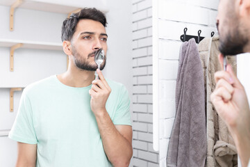 Handsome man doing facial workout with spoon exercises in front of the mirror. Young guy holding...