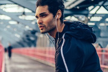Motivated fit man dressed in stylish active clothes listening music payer in earphones and doing warming up before run.Athletic male jogger doing stretching on bridge before workout