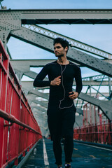 Motivated male jogger dressed in black active wear running and listening music player in earphones enjoying sport lifestyle.Sportsman in tracksuit training in urban setting on bridge