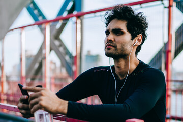 Thougtful young athletic man in earphones listening motivated audio songs via telephone player after workout in urban setting.Professional sportsman in active wear standing on bridge during break