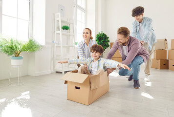 Happy family couple with children on moving day having fun in new apartment riding son in cardboard...