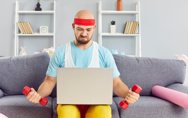 Funny young man in bright sportswear using dumbbells exercising following trainer online via laptop...
