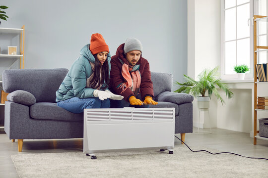 Full length photo of a young frozen couple sitting on the sofa in the living room in winter outerwear and hats at home and trying to warm their hands on electric heater. Heating problems concept.