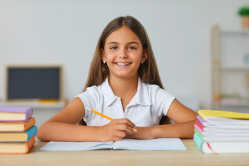 Portrait of a smiling happy schoolchild girl sitting at the desk in classroom looking cheerful at...