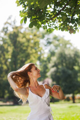 A carefree young woman with long hair experiences the joy of a sunny day in a lush green park, embracing a gentle summer breeze.