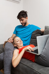 A young man and woman enjoy a relaxed moment on a sofa, as the woman lies down with a laptop and the man looks on with affection.