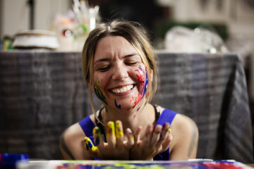 A happy female painter with vibrant paint smeared on her face and hands enjoys a fun moment in her...
