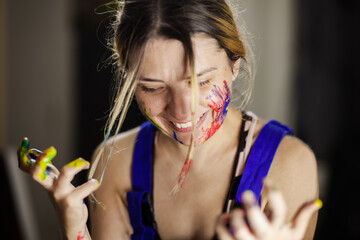 A happy young woman artist laughs as she is splattered with vibrant colored paint, showcasing...