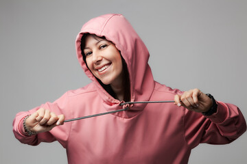 Smiling young woman adjusting her pink hoodie on a gray background