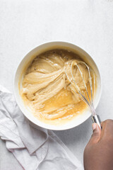 Overhead view of vanilla cake batter being whisked in a white ceramic bowl, flatlay of cake batter being mixed, process of making cake